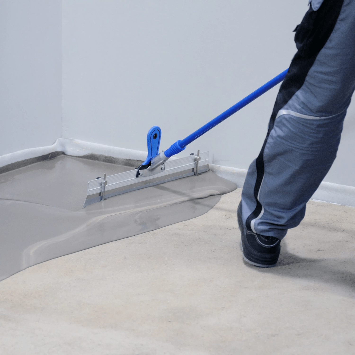 Worker spreading self-leveling underlayment with a blue-handled gauge rake across an indoor floor